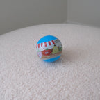 A small round blue plastic toy capsule or ball with colorful cartoon graphics and text, resting on a textured white fabric surface against a plain light gray background.