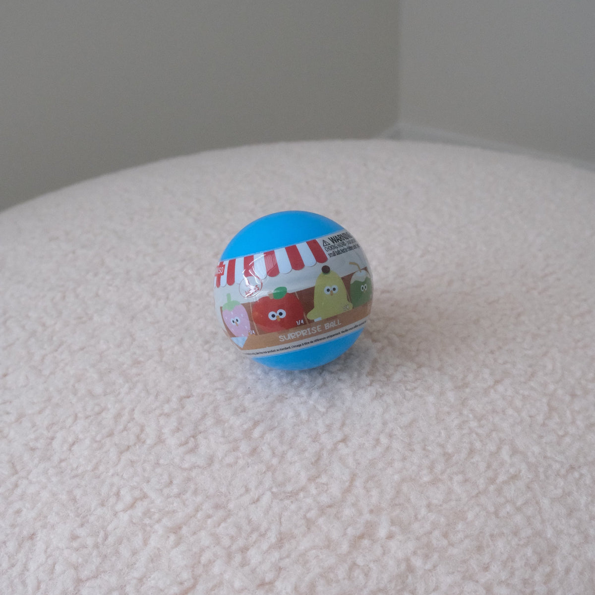 A small round blue plastic toy capsule or ball with colorful cartoon graphics and text, resting on a textured white fabric surface against a plain light gray background.
