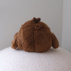 A brown plush stuffed animal with floppy ears and a small dark bow on top, photographed from behind on a white textured surface against a plain light gray wall.
