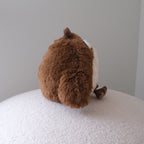 A brown plush toy with a white belly, photographed from behind while sitting on a white textured surface against a plain light gray wall.