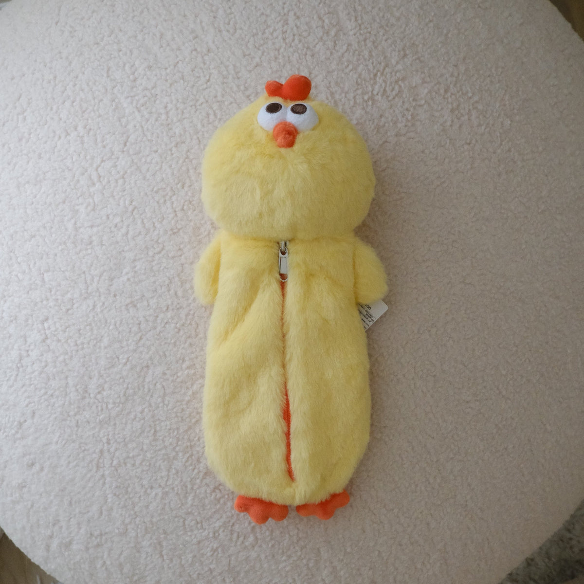 A yellow plush chicken pencil case with orange beak and feet, lying on a round cream-colored textured ottoman.