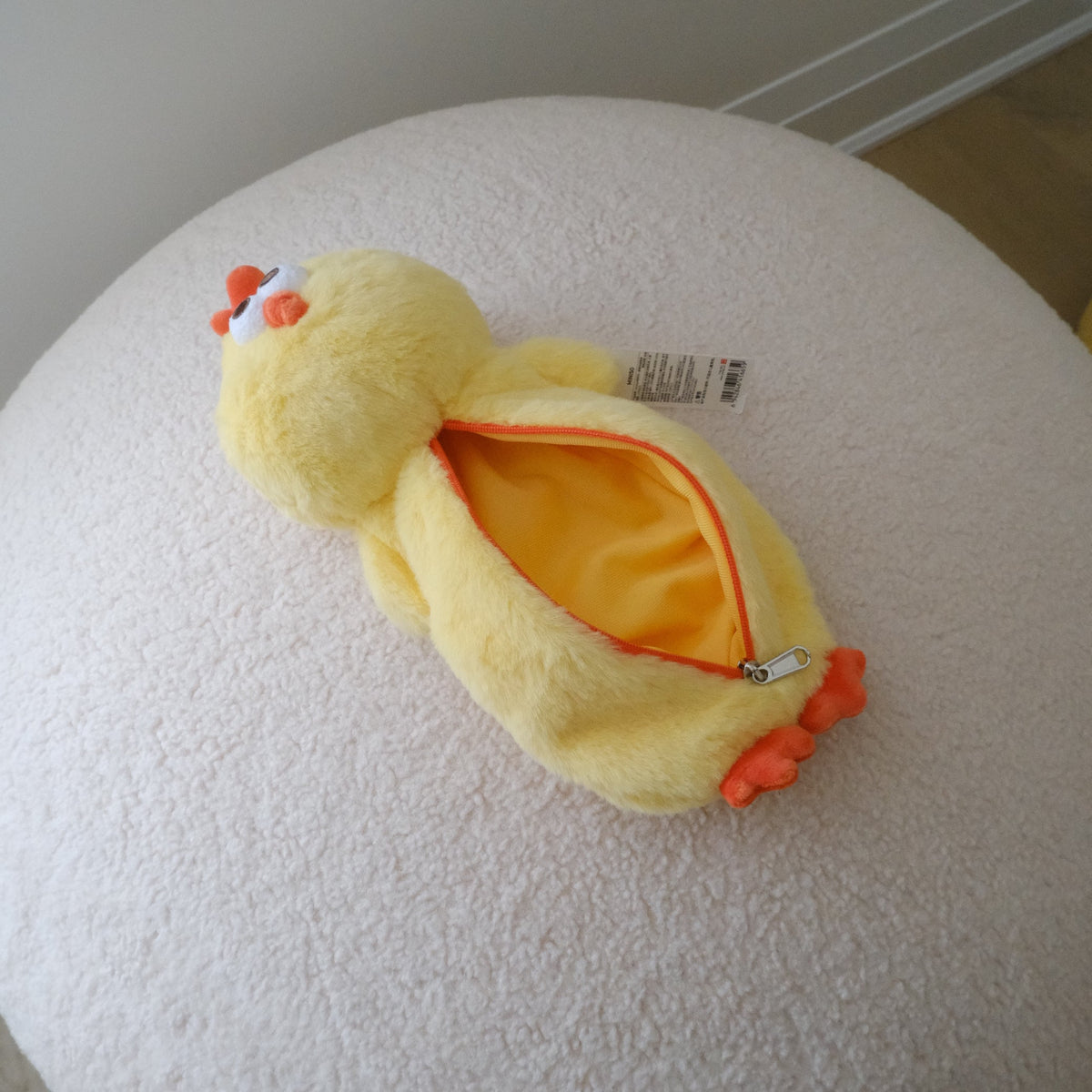 A yellow plush chicken-shaped zippered pouch with orange details, resting on a round white textured ottoman in an indoor setting.