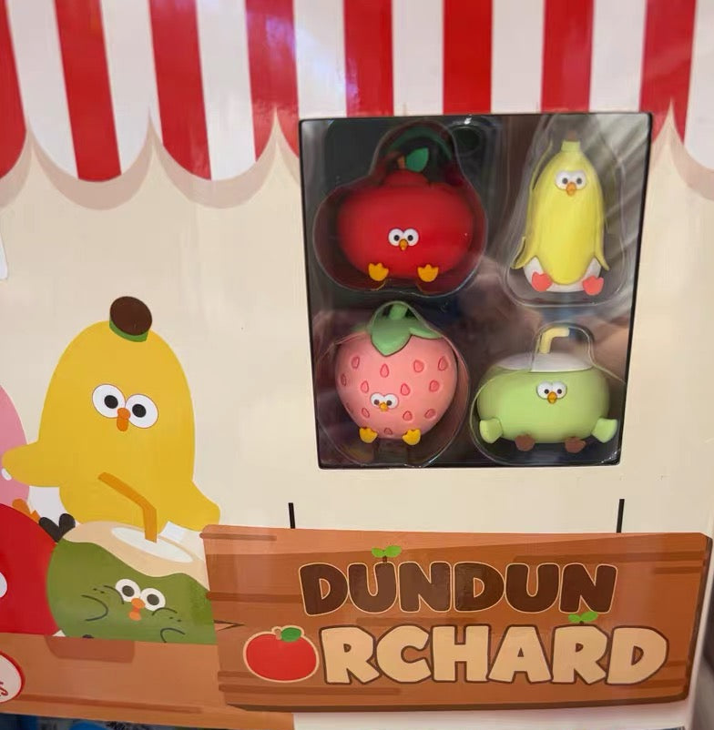 A display case filled with blue and red plastic surprise eggs, with a large window above showing four small fruit-shaped plush toys labeled 'DUNDUN ORCHARD'.