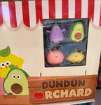 A retail display box for Dundun Orchard surprise eggs, showing four small avocado-themed squishy toys in a clear window. The display box has a cream background with red-and-white striped awning graphics and cartoon avocado illustrations.