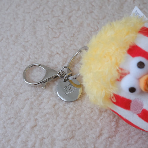 A small plush chicken keychain with a silver lobster clasp and round metal tag, lying on a textured cream fabric surface. The plush has yellow fluffy feathers, red and white stripes, and a white face with black eyes.