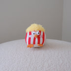 A small plush bird toy with red and white vertical stripes, yellow fluffy hair, and orange feet, sitting on a textured white surface against a plain light gray background.