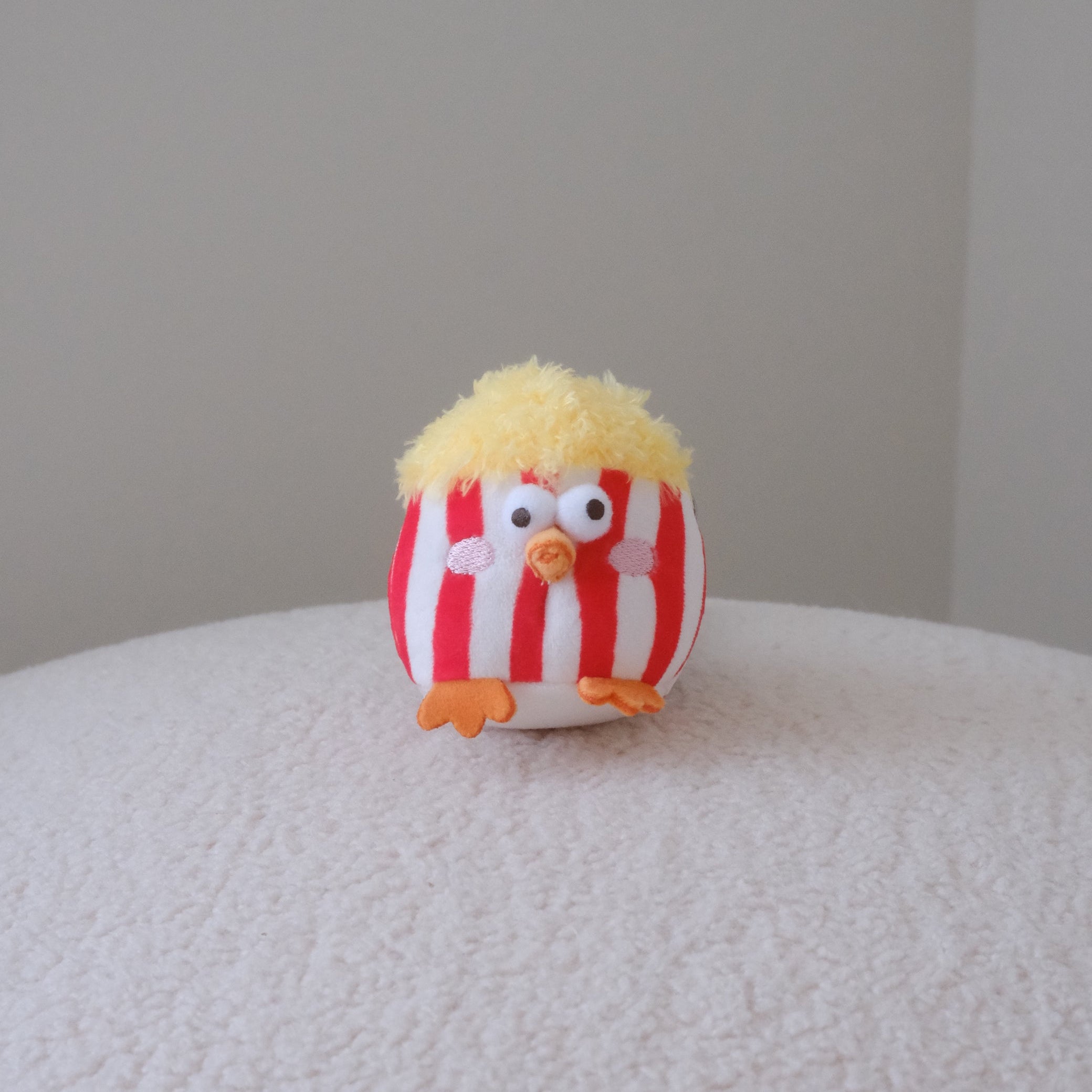 A small plush bird toy with red and white vertical stripes, yellow fluffy hair, and orange feet, sitting on a textured white surface against a plain light gray background.