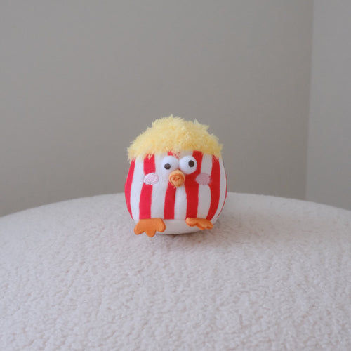 A small plush bird toy with red and white vertical stripes, yellow fluffy hair, and orange feet, sitting on a textured white surface against a plain light gray background.