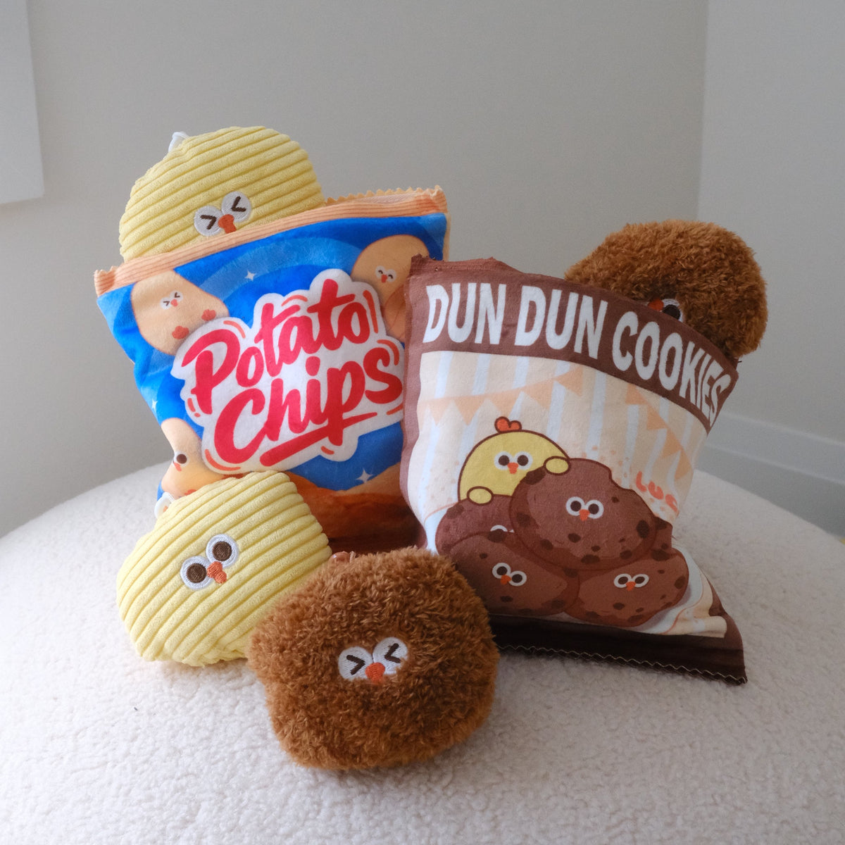 Plush toy snacks styled like Potato Chips and DUN DUN COOKIES bags, arranged on a white textured surface against a plain light gray wall.