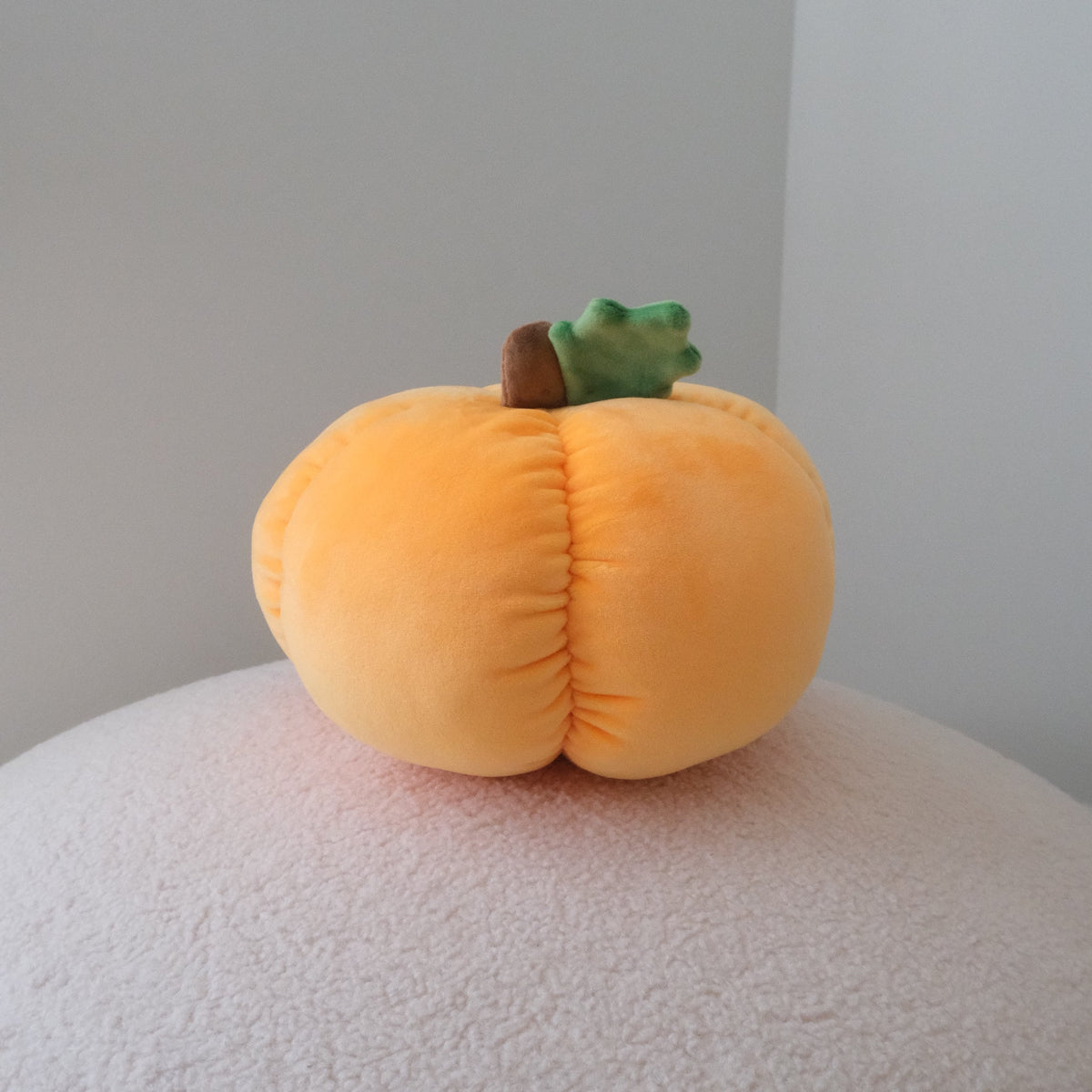 A plush pumpkin-shaped cushion in soft orange-yellow with a small green leaf and brown stem, resting on a textured white surface against a plain light gray wall.