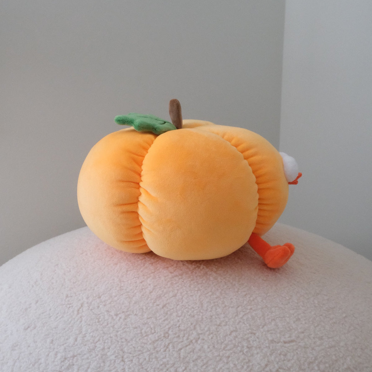 A plush pumpkin toy with a green stem and orange feet, resting on a textured white surface against a plain light gray background.