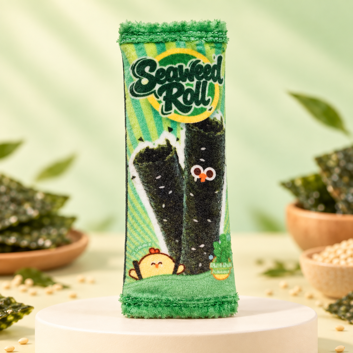A packaged seaweed roll snack stands upright on a white pedestal. The green wrapper features the words 'Seaweed Roll' and an illustration of two dark seaweed rolls, with small decorative props around it including dried seaweed sheets and seeds against a soft green background.