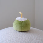 A small plush coconut-shaped toy or cushion in light green and white with a pale yellow straw, standing on a textured white surface against a plain light gray wall.