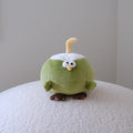 A small plush toy shaped like a green coconut with a white head, yellow curved straw, and brown feet, standing on a white textured surface against a plain light gray wall.