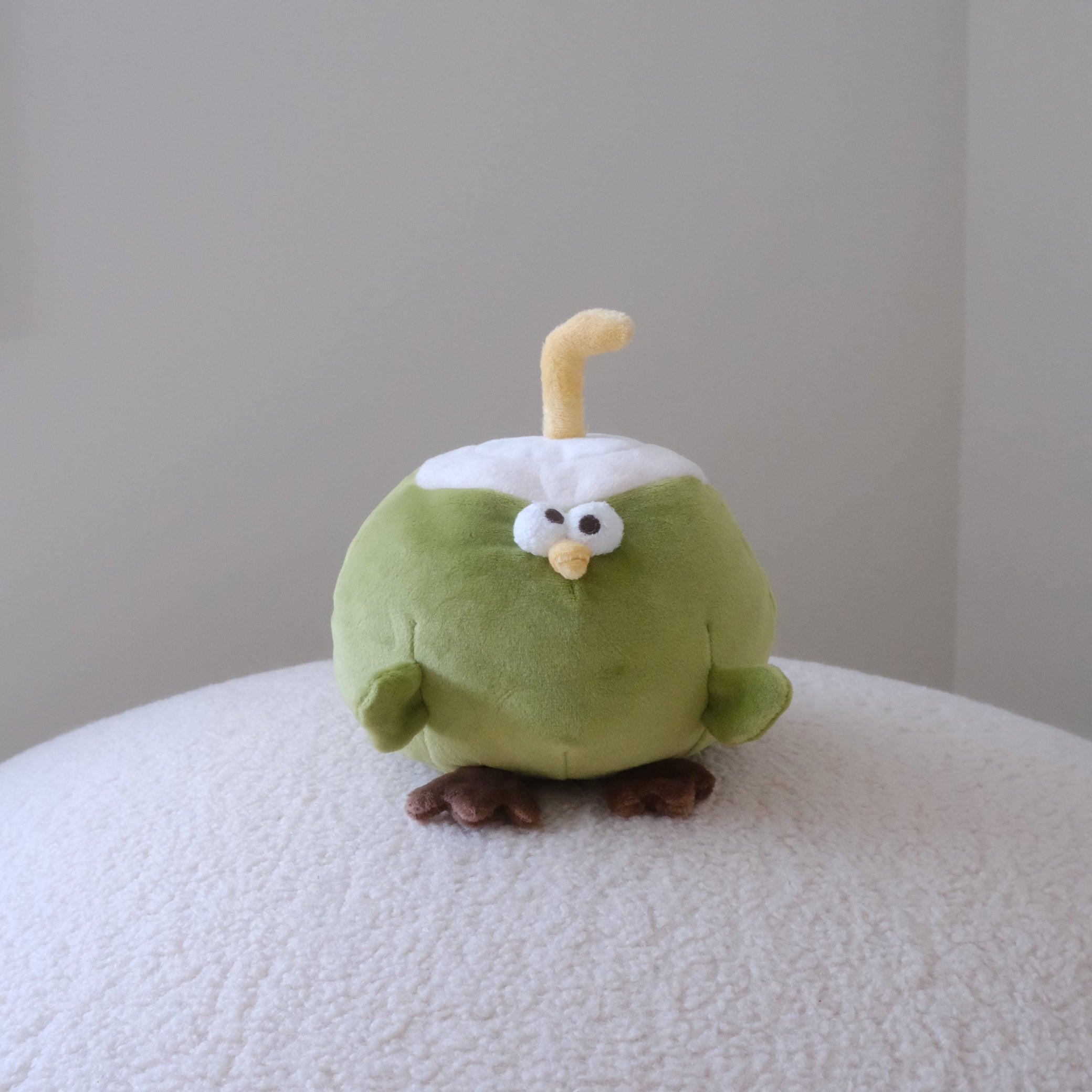 A small plush toy shaped like a green coconut with a white head, yellow curved straw, and brown feet, standing on a white textured surface against a plain light gray wall.