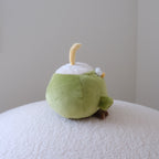 A small plush toy shaped like a green coconut with a white head, yellow curved straw, and small brown detail, sitting on a textured white surface against a plain light gray wall.