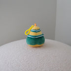 A small plush toy shaped like a striped hat, featuring yellow and green stripes with a yellow loop, resting on a textured white surface against a neutral gray background.