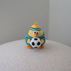 A small clay figurine of a yellow chick wearing a green and white striped sweater, holding a black and white soccer ball, sitting on a textured white surface against a plain gray background.