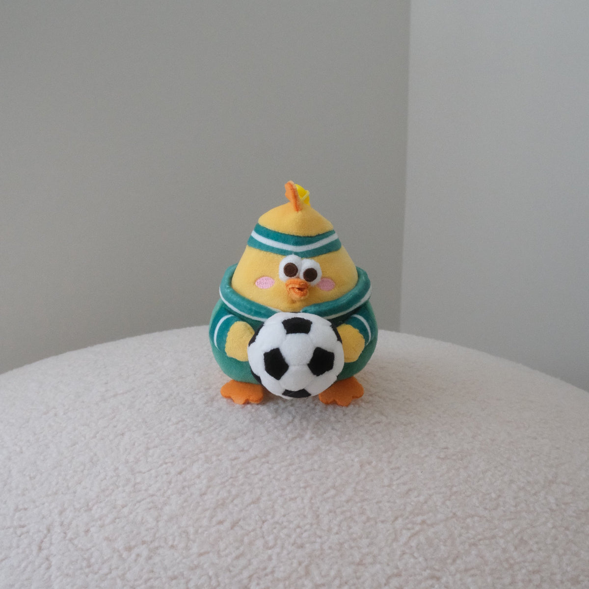 A small clay figurine of a yellow chick wearing a green and white striped sweater, holding a black and white soccer ball, sitting on a textured white surface against a plain gray background.