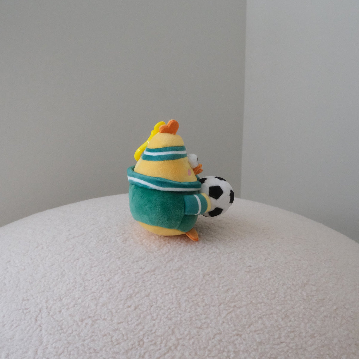 A small plush toy bird wearing a teal and white striped shirt, sitting on a textured white surface and holding a black and white soccer ball, photographed in a minimalist room corner.
