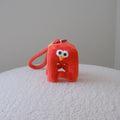 A small red plush character keychain or bag charm with a loop handle, photographed standing on a white textured surface against a plain light gray wall.