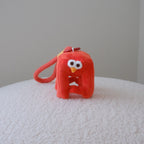 A small red plush character keychain or bag charm with a loop handle, photographed standing on a white textured surface against a plain light gray wall.