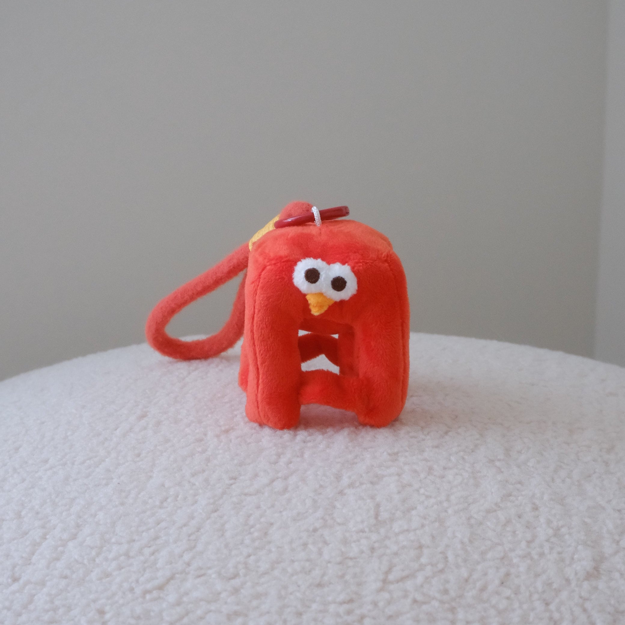 A small red plush character keychain or bag charm with a loop handle, photographed standing on a white textured surface against a plain light gray wall.