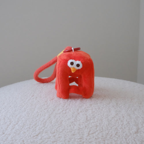 A small red plush character keychain or bag charm with a loop handle, photographed standing on a white textured surface against a plain light gray wall.