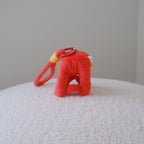 A small red plush toy or accessory with a loop handle and a yellow accent, placed on a white textured surface against a plain light gray wall.