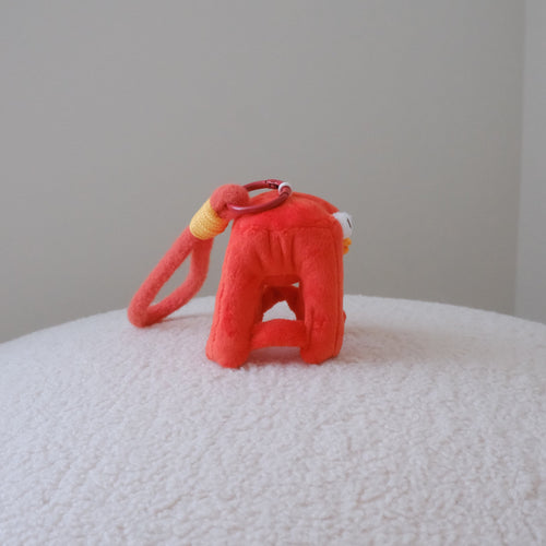 A small red plush toy or accessory with a loop handle and a yellow accent, placed on a white textured surface against a plain light gray wall.