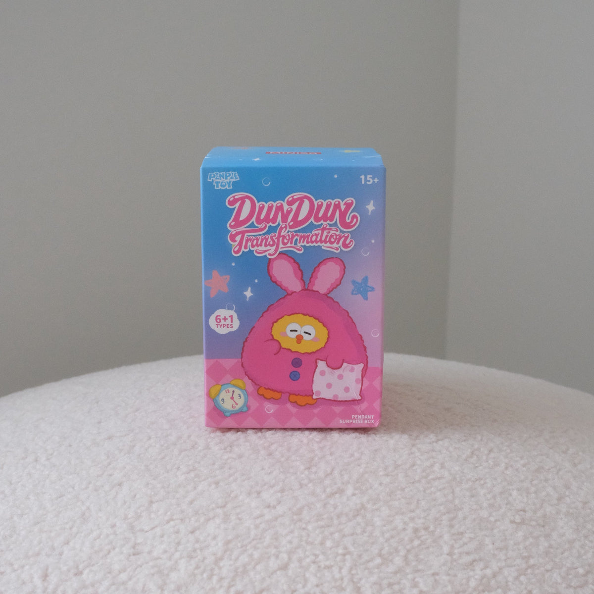 A small blue and pink box of Dundun Transformation mystery packs, featuring a cartoon bunny character, standing on a white textured surface against a plain gray wall.