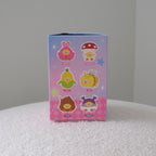 A small rectangular blind-box package standing upright on a white textured surface against a plain light gray wall. The box is pink and blue with six cartoon character illustrations on the front.