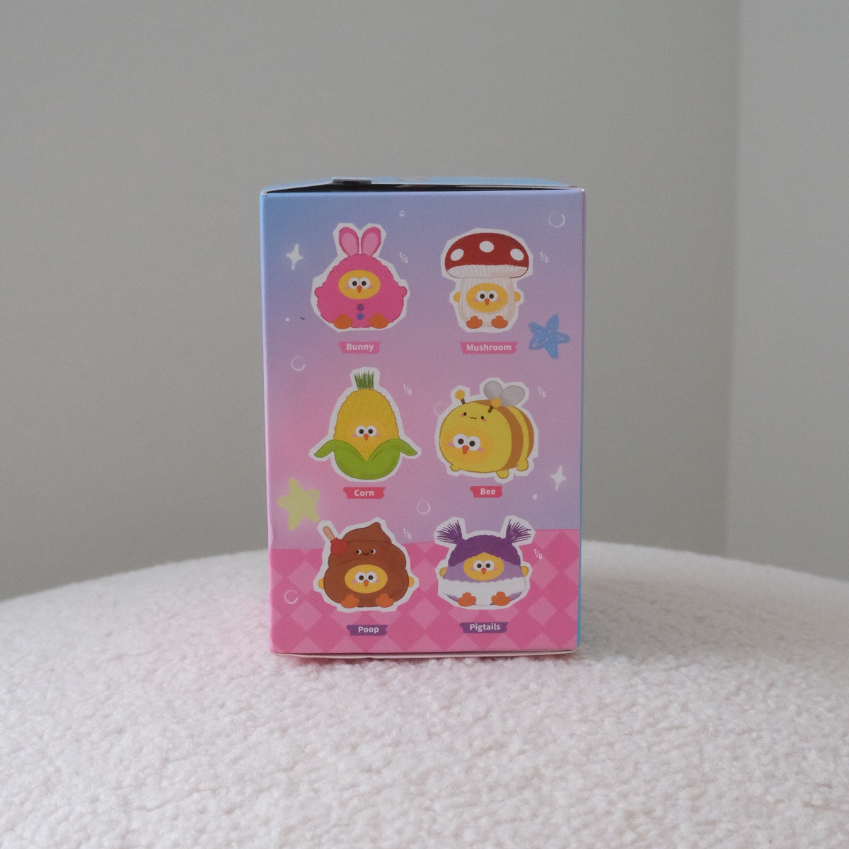 A small rectangular blind-box package standing upright on a white textured surface against a plain light gray wall. The box is pink and blue with six cartoon character illustrations on the front.