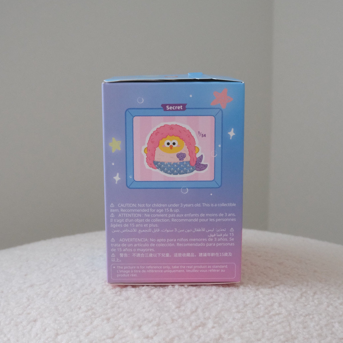 A collectible blind box featuring a cute mermaid illustration with pink hair and a blue tail, standing on a white textured surface against a neutral gray background.
