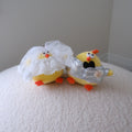 Two small yellow chick-shaped novelty items on a white textured surface against a plain light gray wall. One chick wears a white lace veil, and the other wears a silver foil suit with a black bow tie.