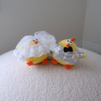 Two small yellow chick-shaped novelty items on a white textured surface against a plain light gray wall. One chick wears a white lace veil, and the other wears a silver foil suit with a black bow tie.
