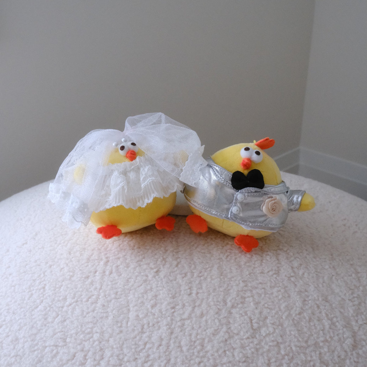 Two small yellow chick-shaped novelty items on a white textured surface against a plain light gray wall. One chick wears a white lace veil, and the other wears a silver foil suit with a black bow tie.