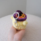 A small plush character keychain held in a hand. The plush is pale yellow with a purple witch hat and orange pumpkin accent, attached to a silver metal key ring.
