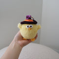 A small yellow plush chick toy with a purple and orange witch hat, held in a person's hand against a plain indoor background.