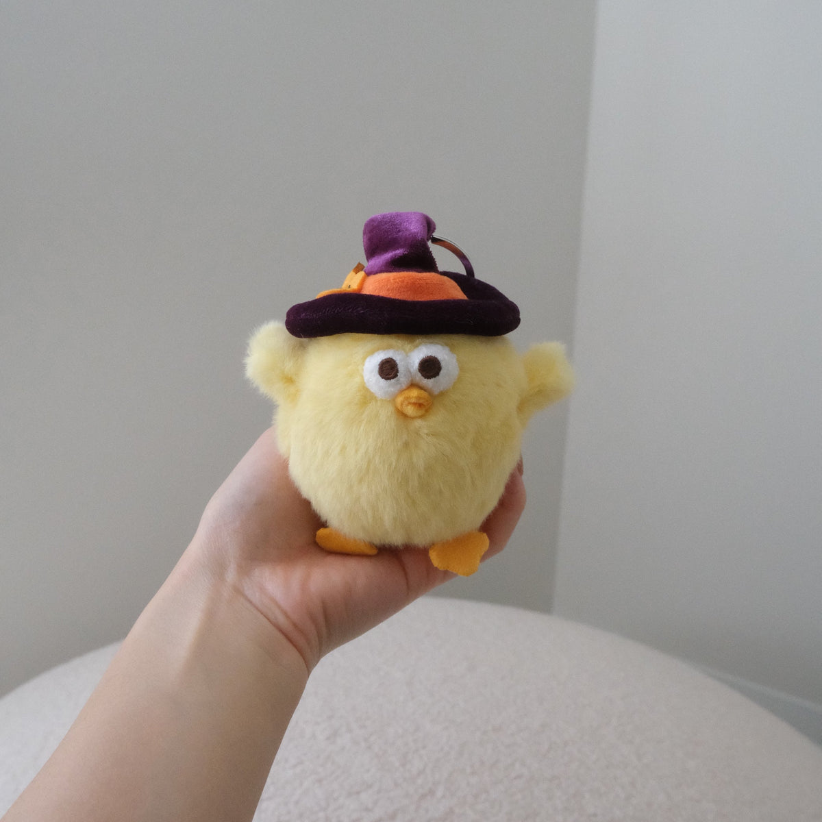 A small yellow plush chick toy with a purple and orange witch hat, held in a person's hand against a plain indoor background.