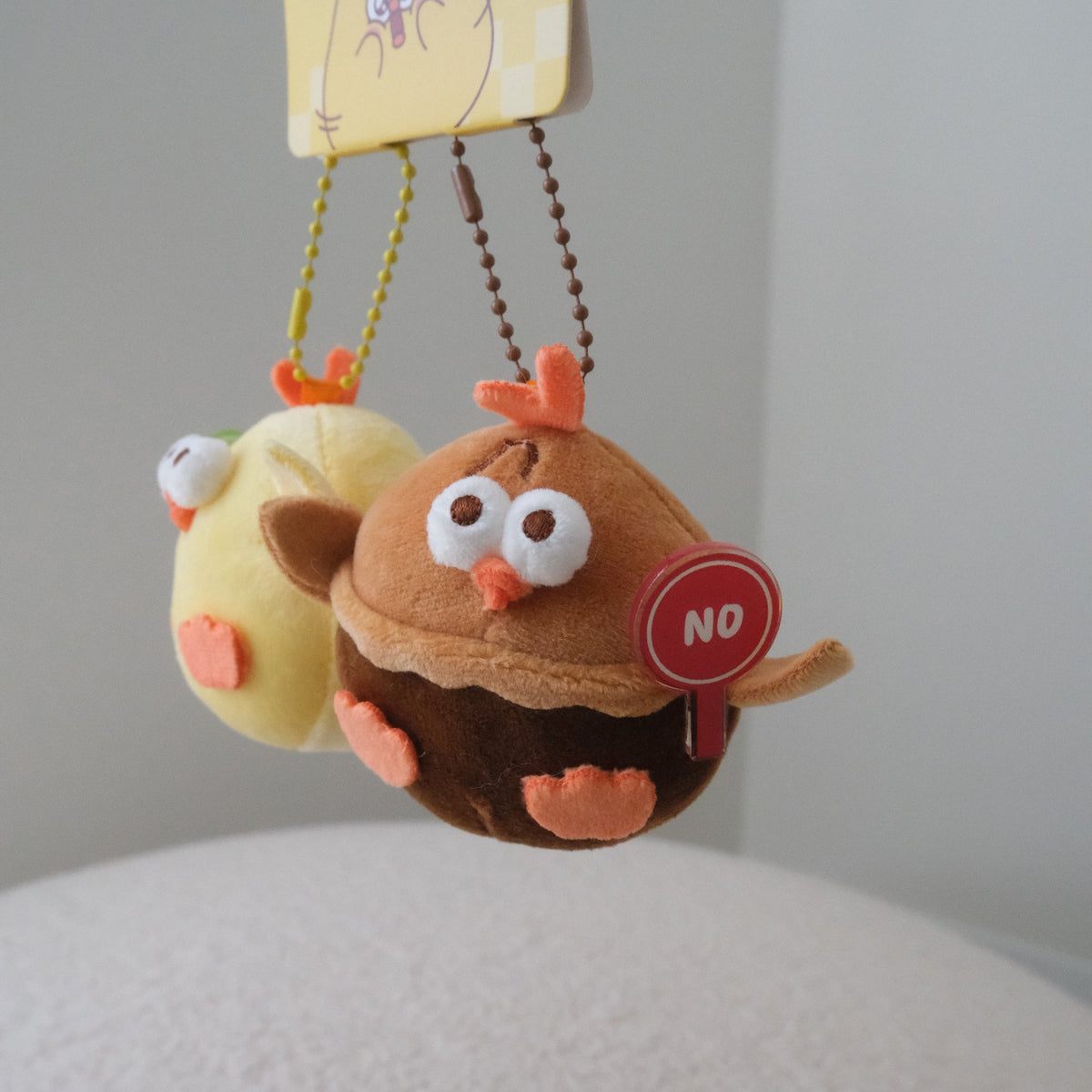 Two cute plush chicken keychains hanging from a yellow card. The front keychain is brown with orange accents and a red 'NO' sign, while the back one is pale yellow.