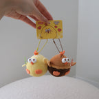 A hand holds a yellow card attached to two small plush character keychains against a plain indoor background. One plush is pale yellow with orange accents, and the other is brown with orange and white details.