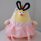 A yellow plush chick toy wearing a pink dress photographed indoors against a plain light gray wall and white textured surface.