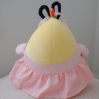 A plush toy or cushion shaped like a round yellow figure with a pink ruffled skirt, pink side details, and black ear-like loops on top, photographed against a plain light gray wall and white textured surface.