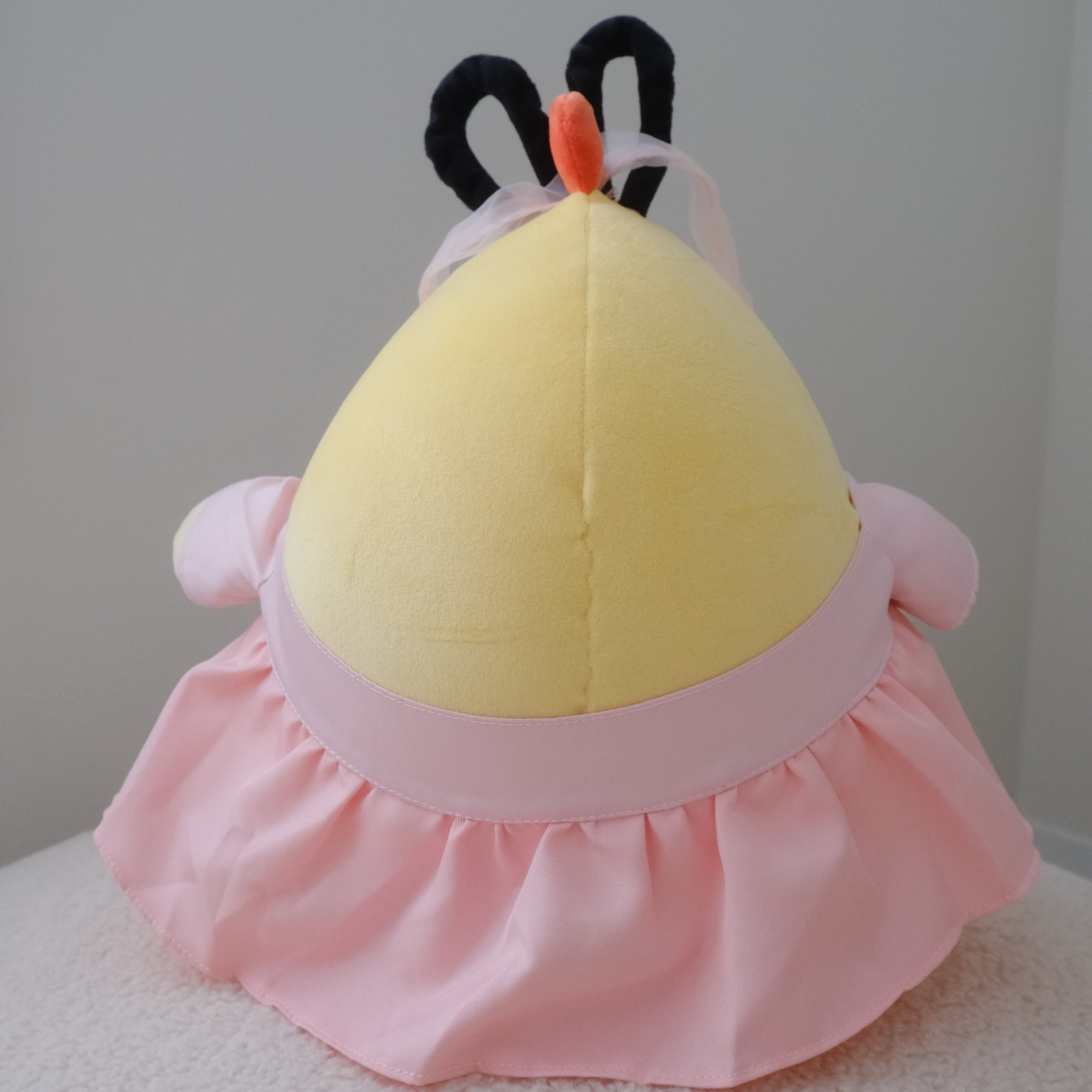 A plush toy or cushion shaped like a round yellow figure with a pink ruffled skirt, pink side details, and black ear-like loops on top, photographed against a plain light gray wall and white textured surface.