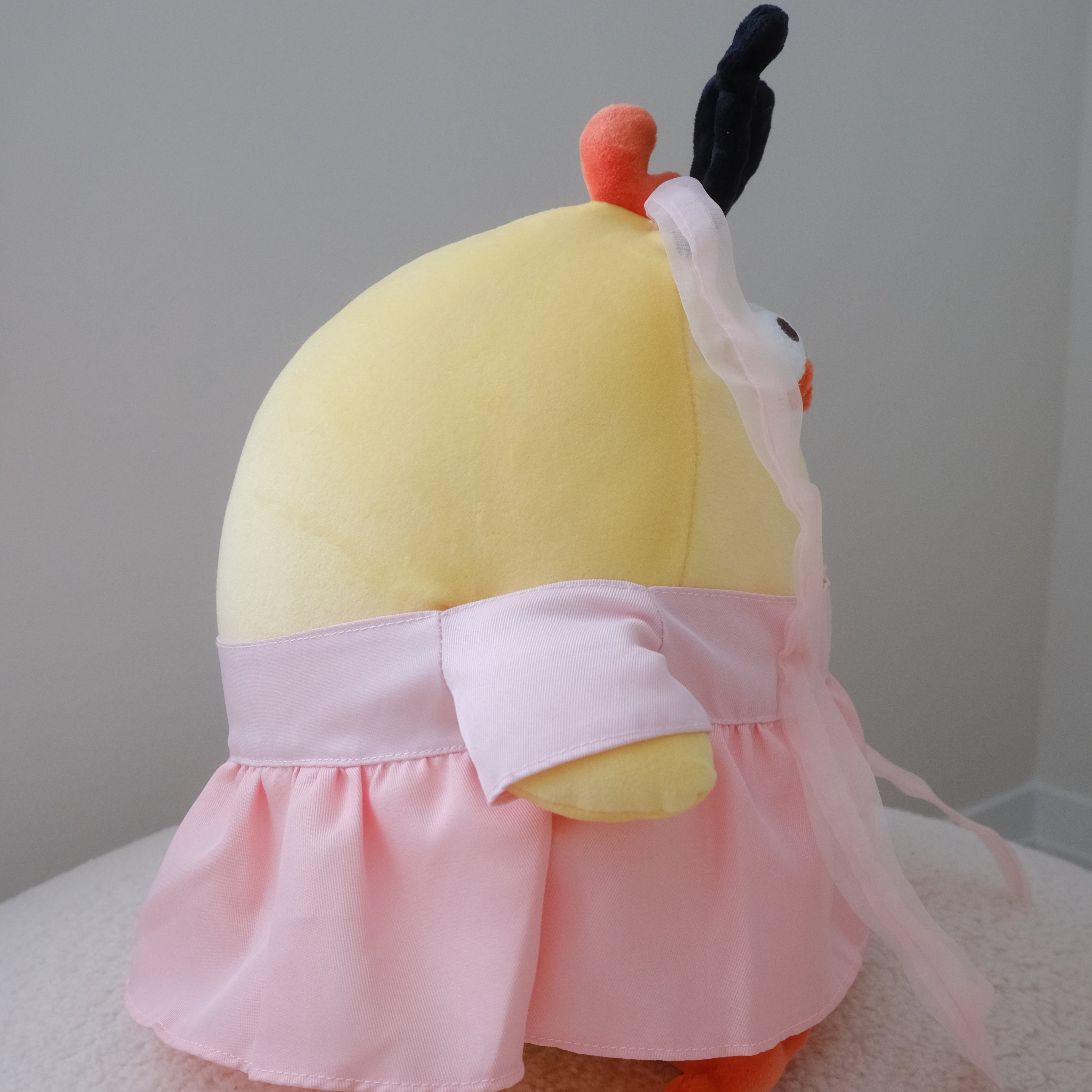 A yellow plush toy with orange feet wearing a pink ruffled skirt and bow, photographed against a plain light gray wall and white textured surface.