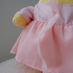 A close-up of a plush toy or doll wearing a light pink dress with a ruffled waistband and sheer pink ribbon, with yellow and orange plush limbs visible against a soft neutral background.
