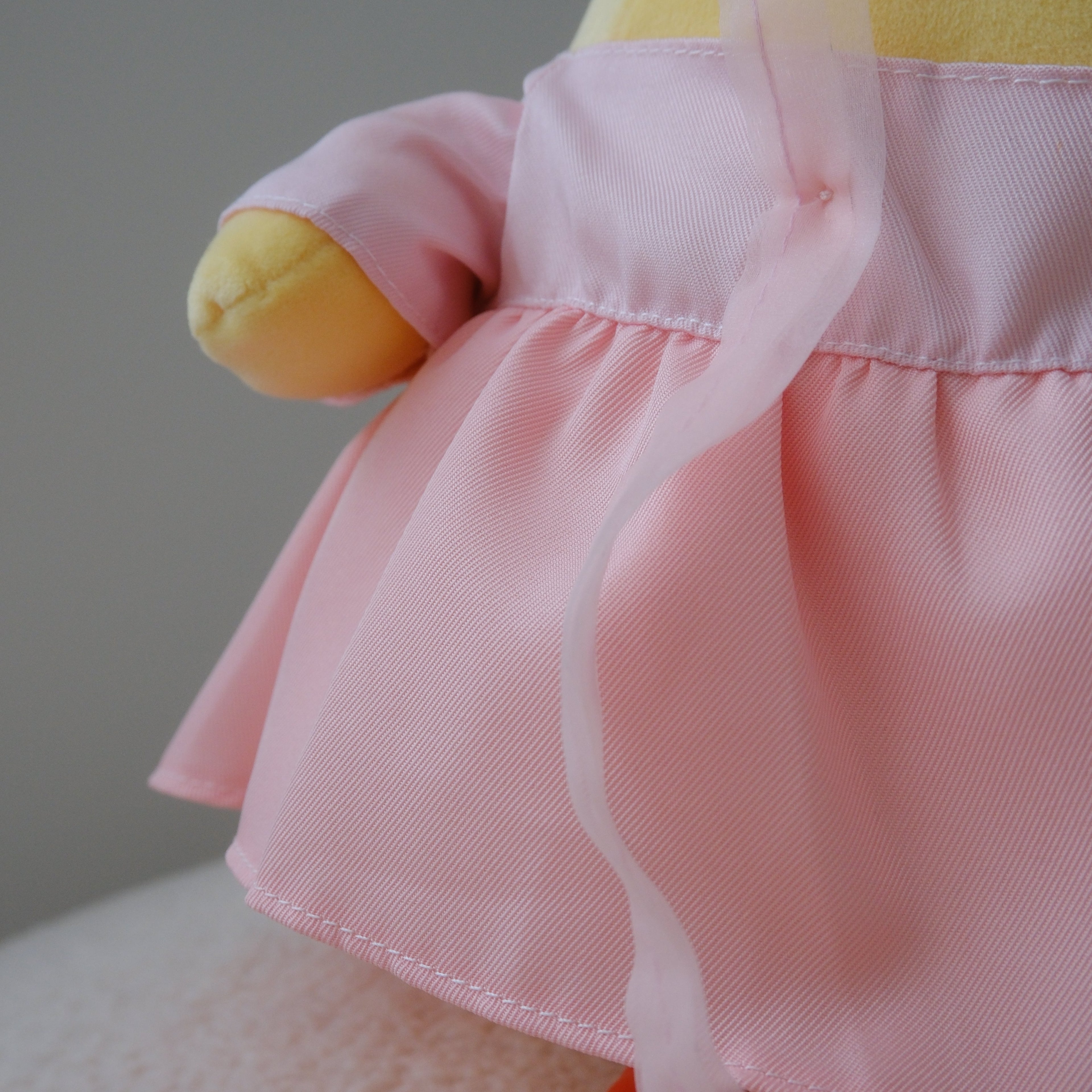 A close-up of a plush toy or doll wearing a light pink dress with a ruffled waistband and sheer pink ribbon, with yellow and orange plush limbs visible against a soft neutral background.