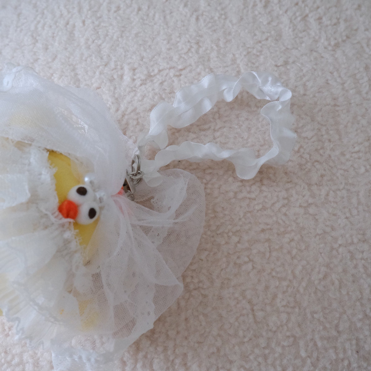 A yellow chicken plush wearing a white tulle wedding dress and attached to a white ruffled wristlet strap, displayed on a textured cream fabric surface.