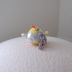 A small yellow plush toy wearing a silver metallic shirt and a ruffled lavender-gray ruffled wristlet strap, resting on a textured cream-colored surface against a plain light gray wall.
